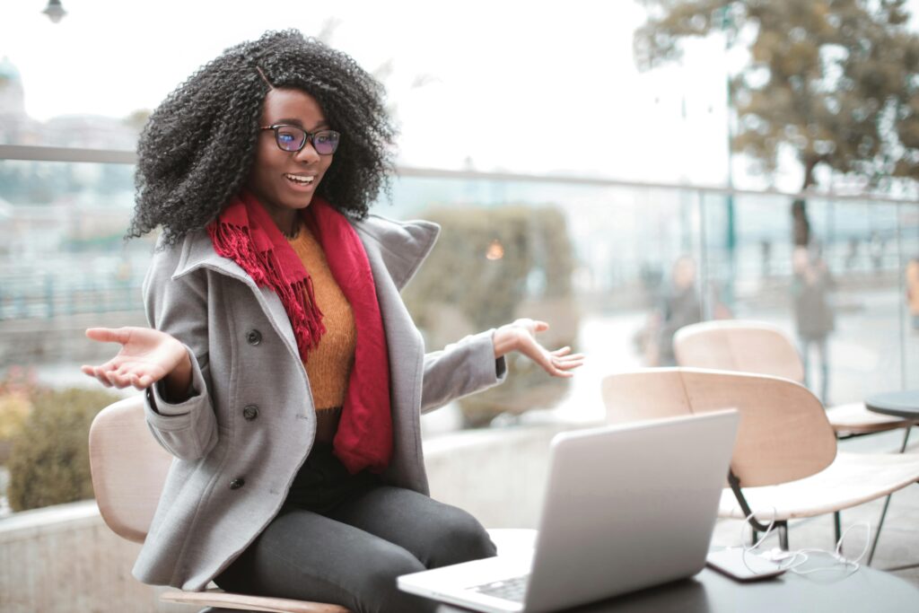 Happy excited African American female laughing and gesticulating while having video calling on laptop and sitting at modern cafe